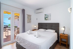 Ein Hotelzimmer mit einem Bett, zwei Nachttischen, einer Klimaanlage, einem Fenster mit Vorhängen, einem Tisch mit Stühlen und Meerblick.