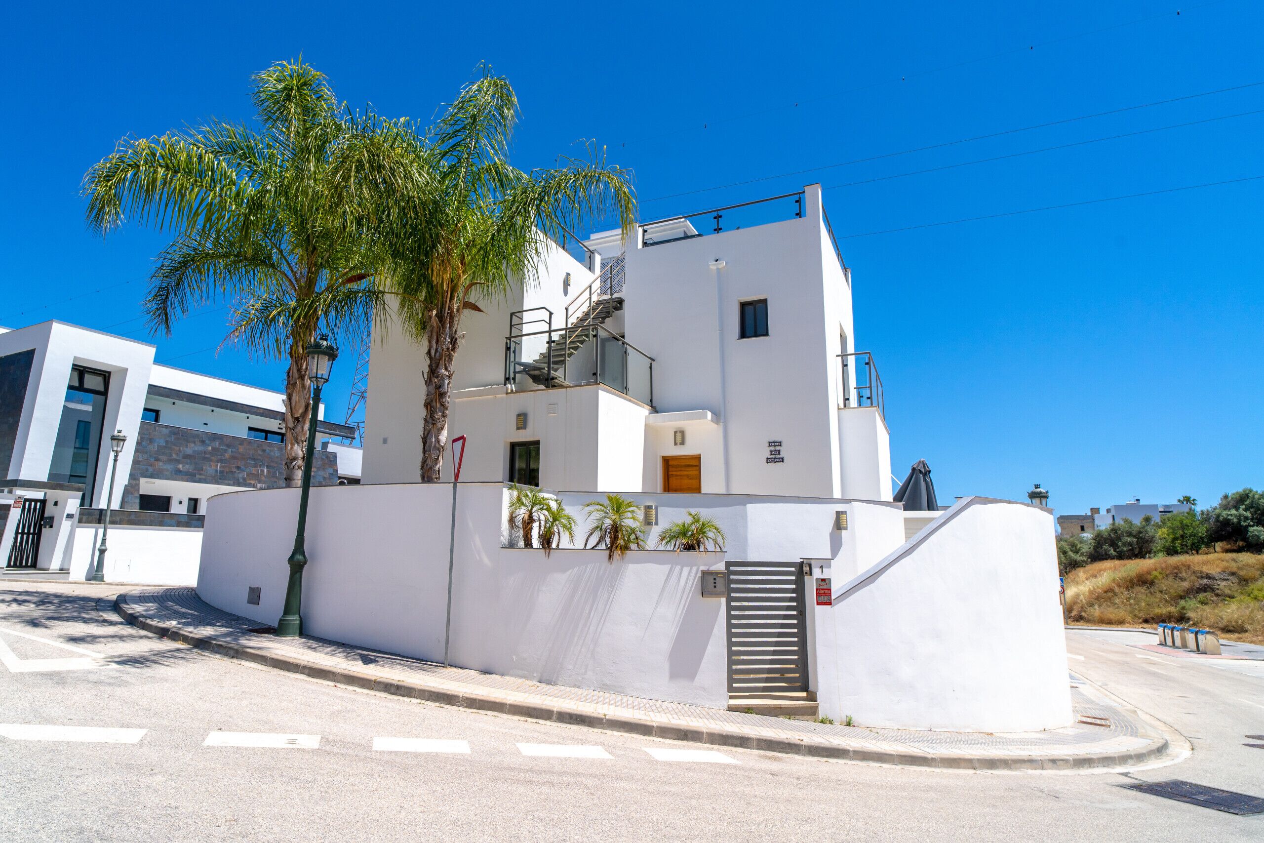 A modern white building with a staircase, palm trees, and a gated entrance.