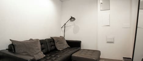 A modern living room with a black leather sofa, two matching cushions, a small ottoman, a floor lamp, and a wall-mounted light switch.