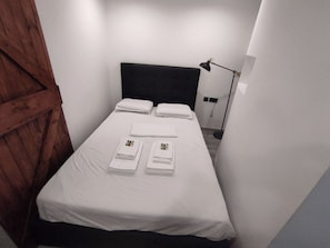 Un lit simple avec du linge de lit blanc et deux serviettes pliées, une tête de lit noire et une lampe de chevet.