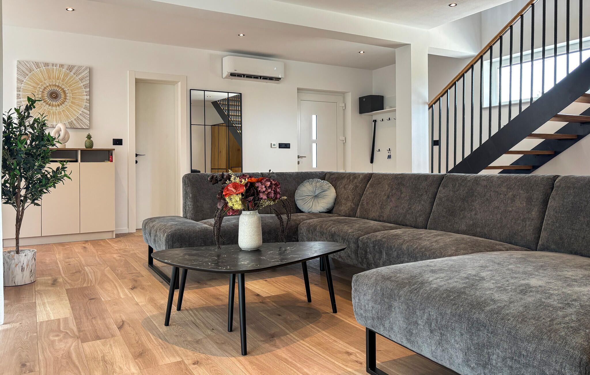 Een moderne woonkamer met een grijze hoekbank, een ronde salontafel en een potplant.