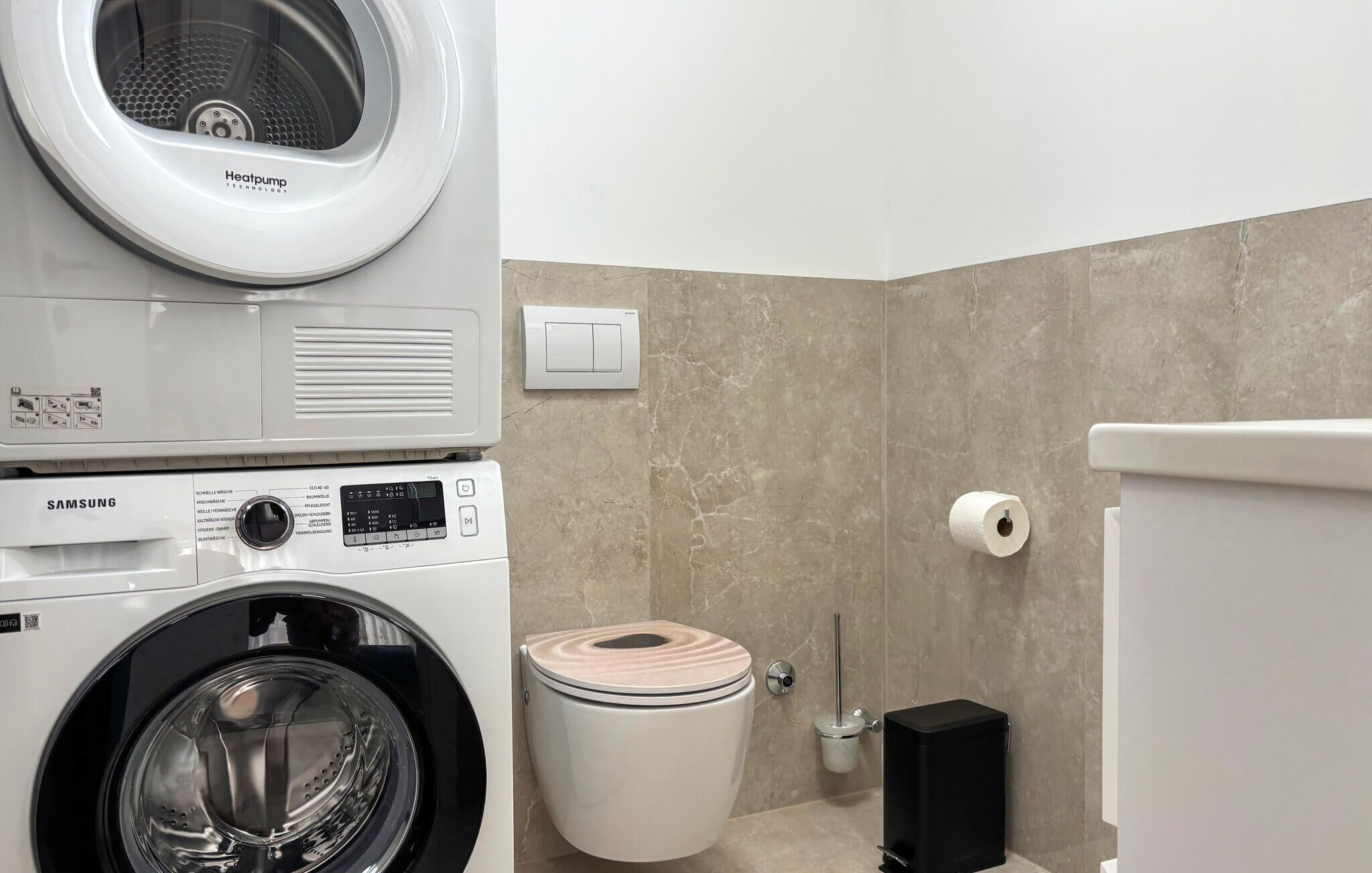 A compact bathroom with a washing machine, toilet, and a trash bin.