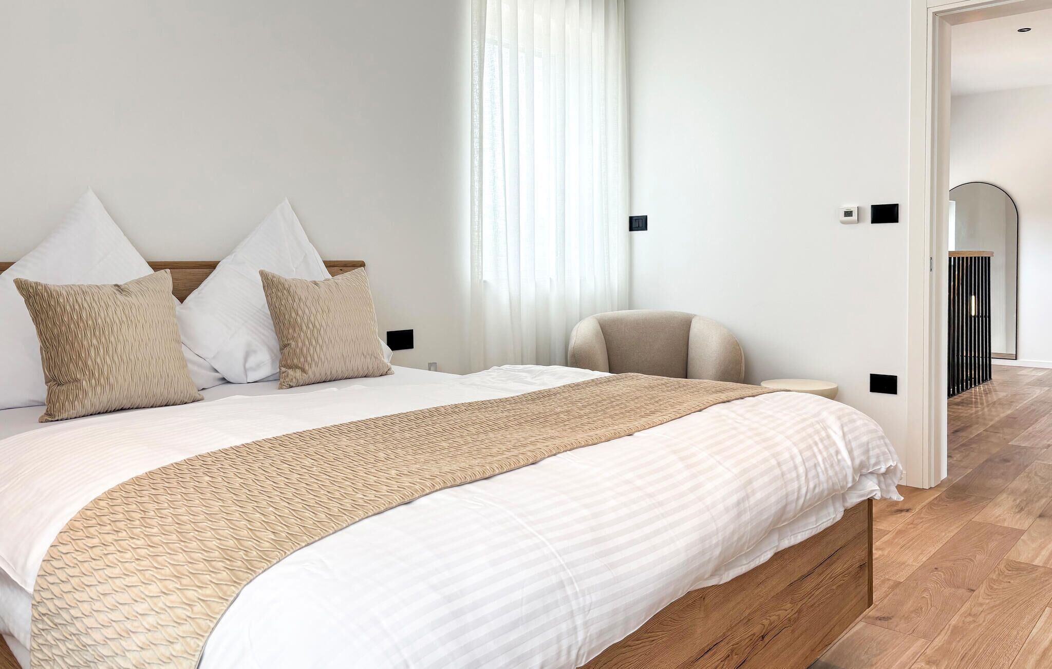 A neatly made bed with a wooden headboard, a bedside chair, and a large window with sheer curtains.