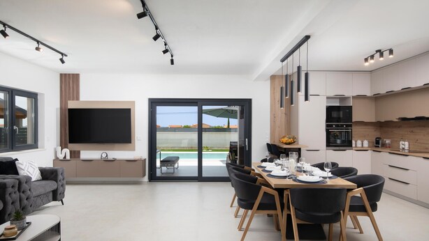Ein modernes Wohnzimmer mit Flachbildfernseher, eine Essecke mit Tisch für vier Personen und eine Küche mit Holzschränken und Backofen.