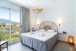 Ein Schlafzimmer mit einem großen Bett, Blick ins Grüne, einem Rattan-Kopfstück und einer Nachttischlampe.