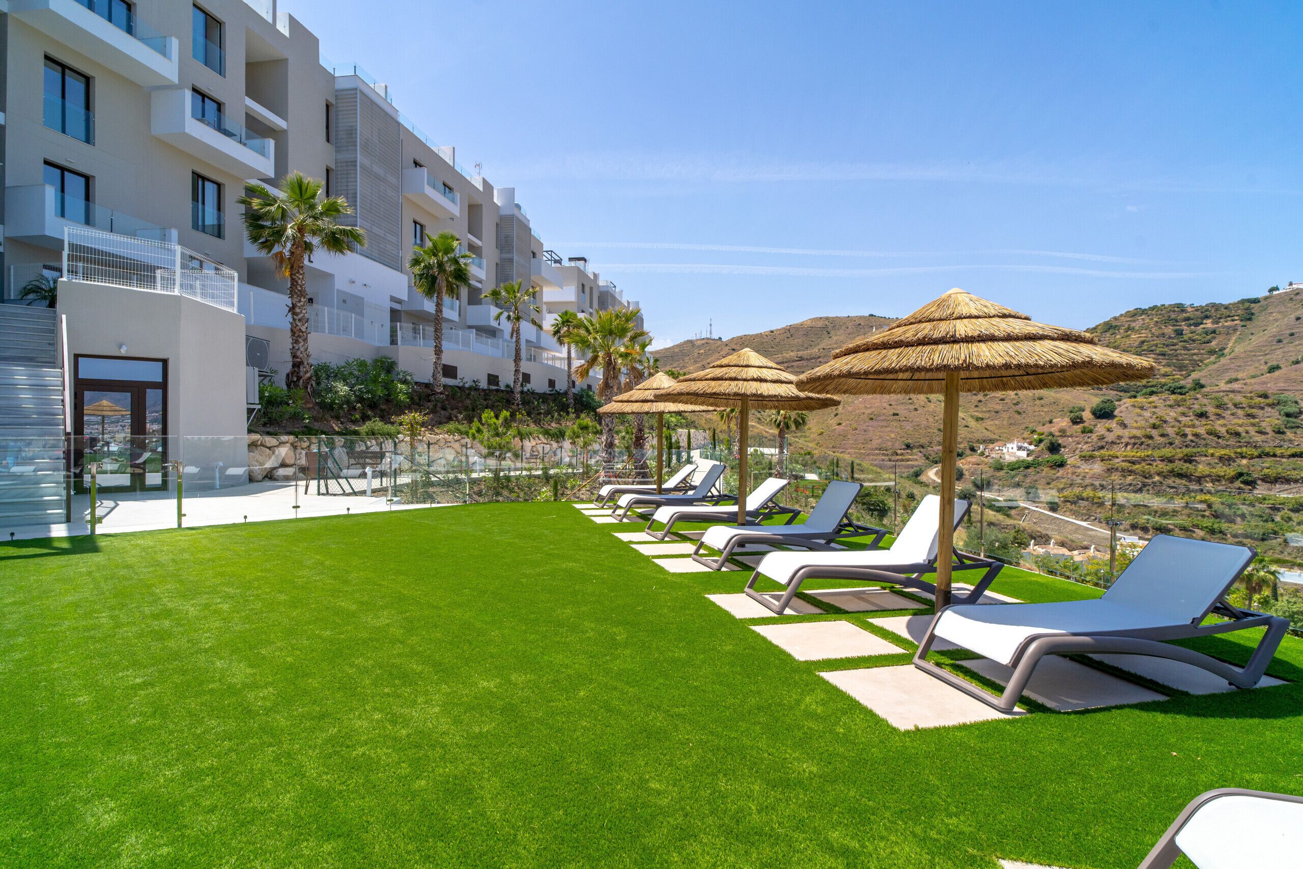 A hotel courtyard with a green lawn, lounge chairs, and thatched umbrellas.