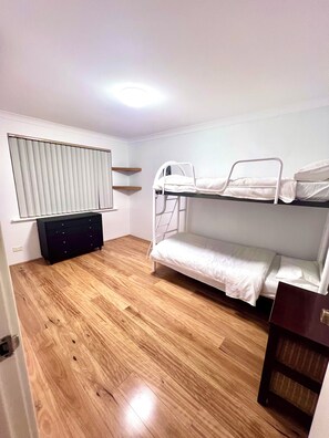 Een kamer met een stapelbed, een dressoir, een raam met jaloezieën en een houten vloer.