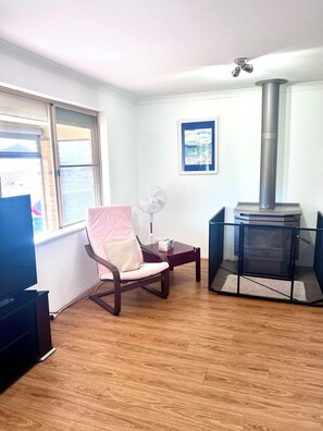 A room with a wooden floor, a pink chair, a table with a fan, a fireplace, and a window.