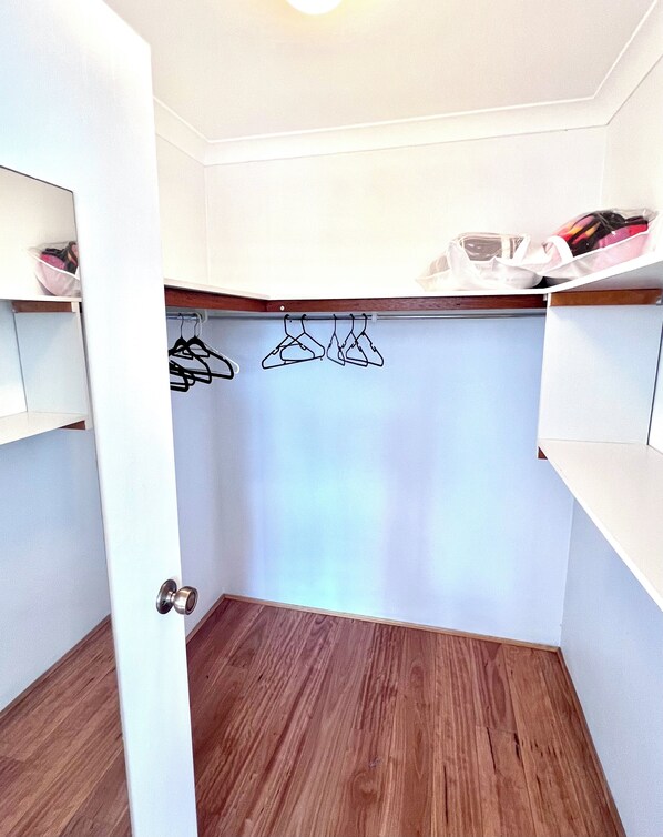 Een inloopkast met houten vloer, witte muren en een kledingroede met kleding.