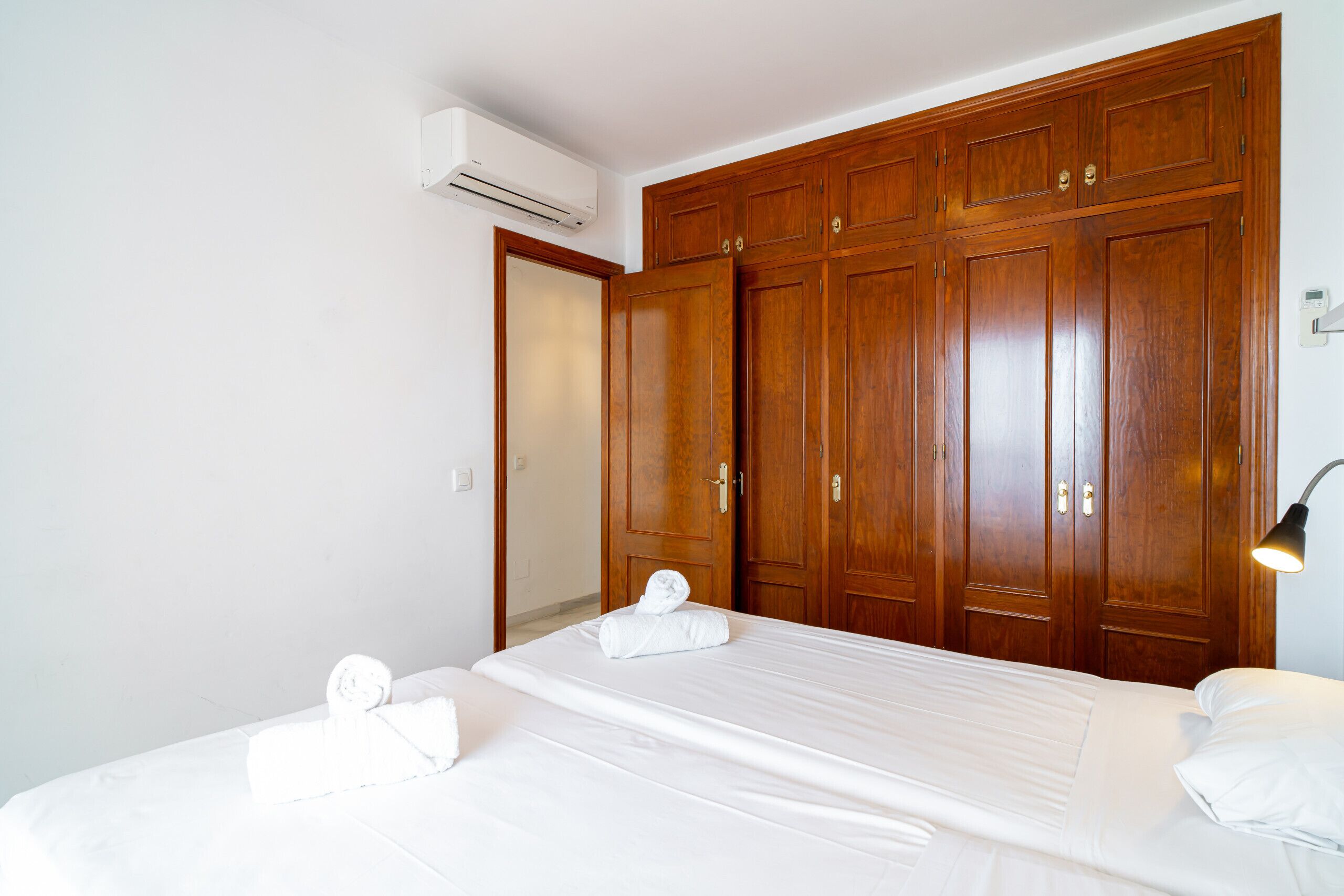 A hotel room with a large wooden wardrobe, a bed with white linens, and a wall-mounted air conditioning unit.
