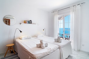 Ein Hotelzimmer mit zwei Betten, einem kleinen Hocker, einem Nachttisch, einem Spiegel und Meerblick durch große Fenster.