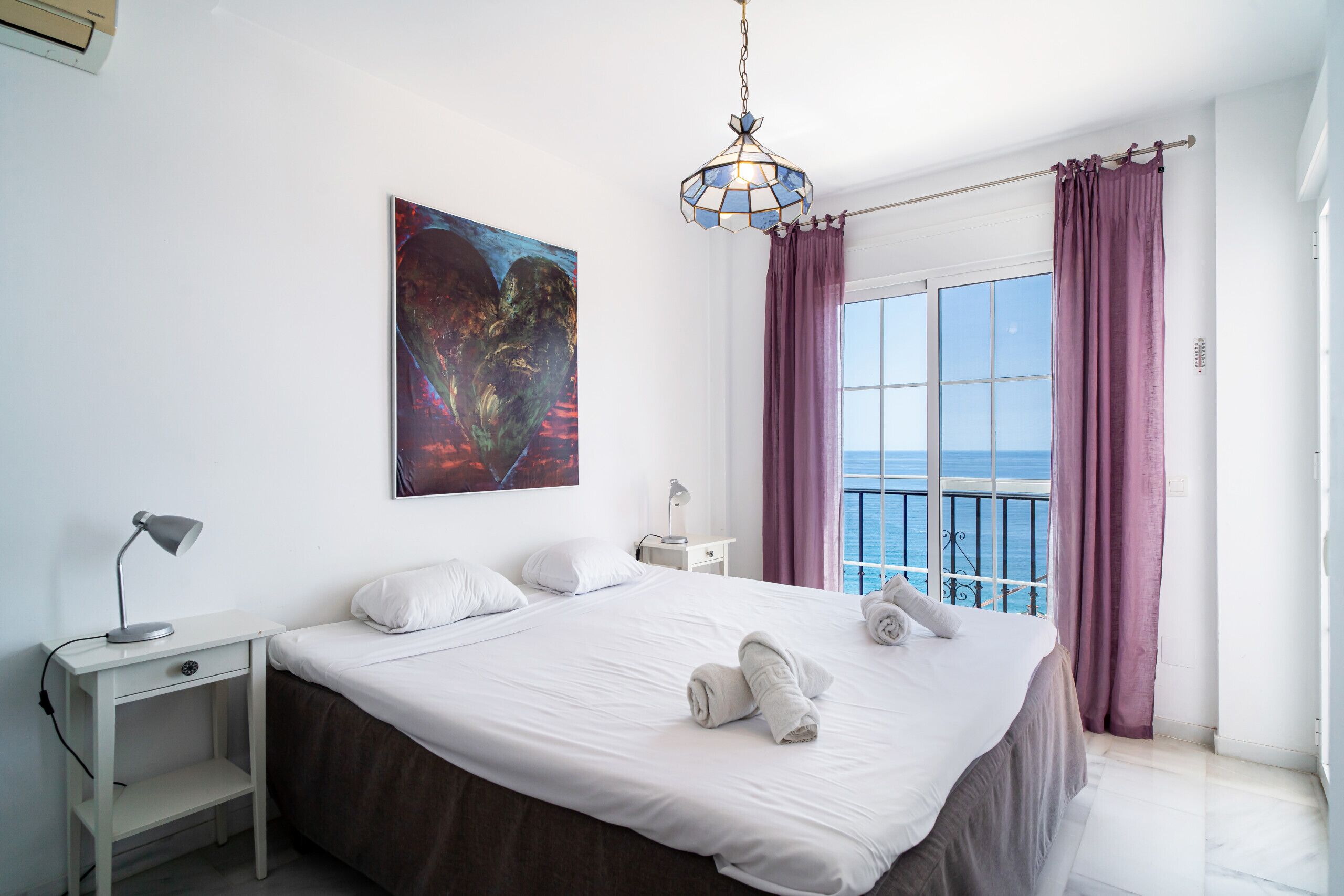 A bedroom with a large bed, a bedside table with a lamp, a painting on the wall, and a view of the sea through a window with curtains.