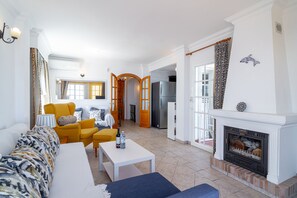 Ein Wohnzimmer mit einer Couch, Sesseln, einem Couchtisch, einem Kamin und einem durch einen offenen Durchgang sichtbaren Küchenbereich.