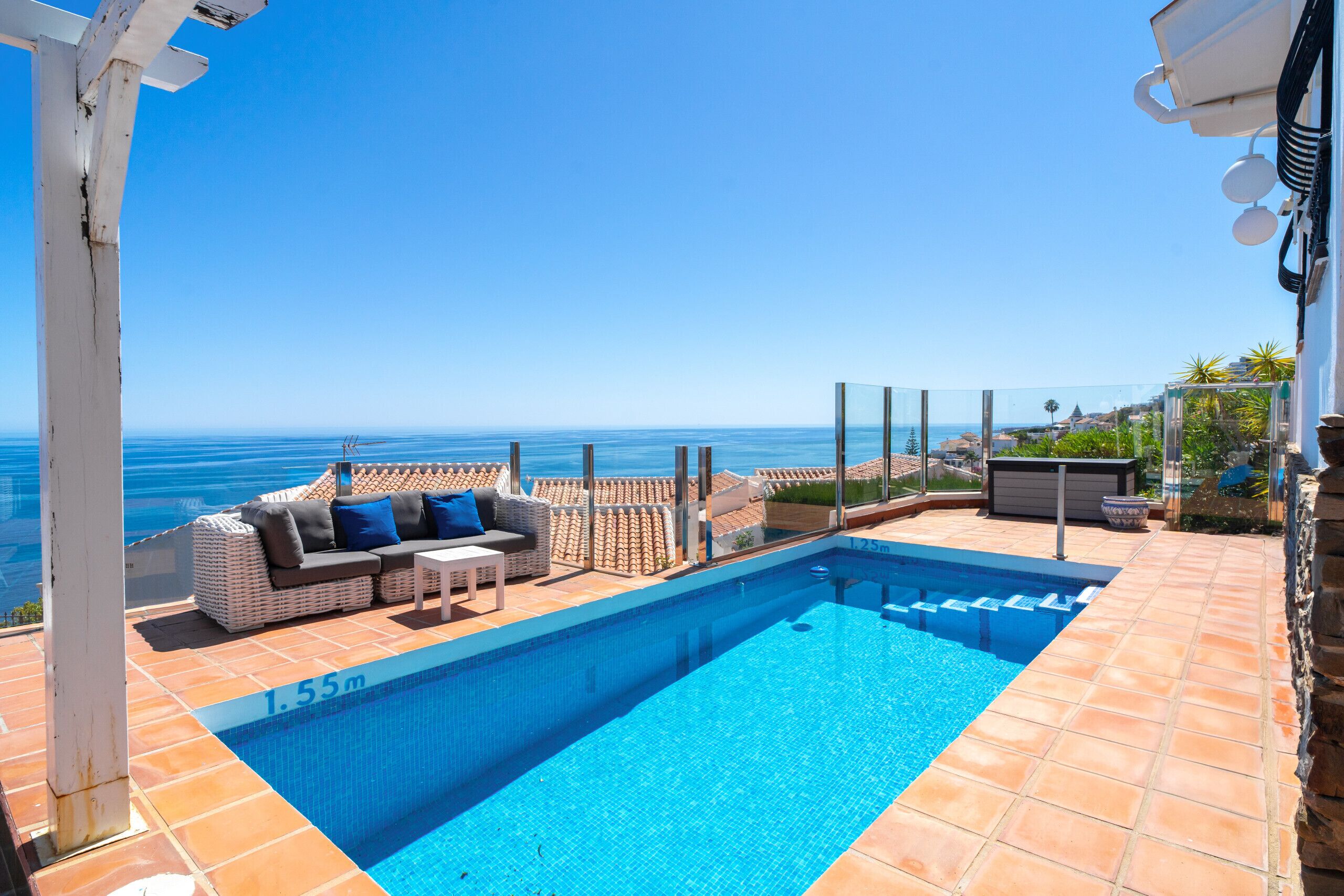 A rooftop pool with a clear view of the sea, a seating area with cushions, and a tiled deck.