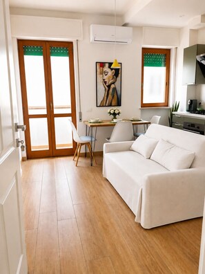 A modern living room with a white sofa, a dining table with chairs, a painting on the wall, and an air conditioning unit. - Elegant apartment 2 km from Poetto (Quartu Sant'Elena)