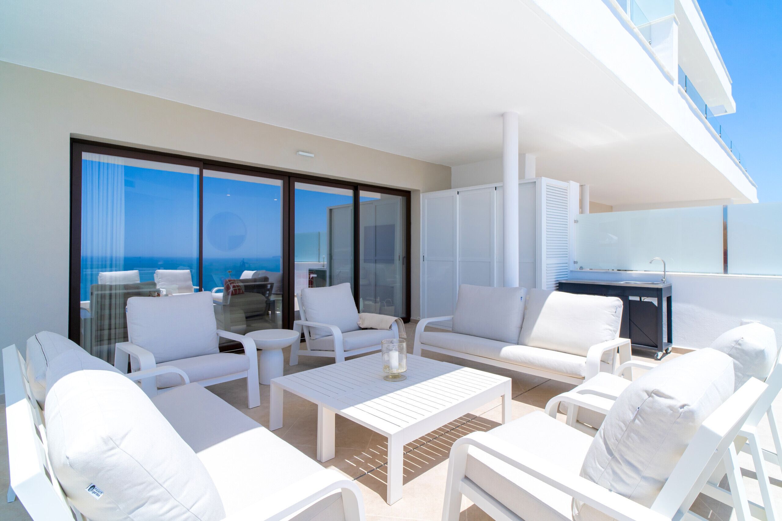 A modern outdoor lounge area with white furniture, large windows, and a view of the sea.