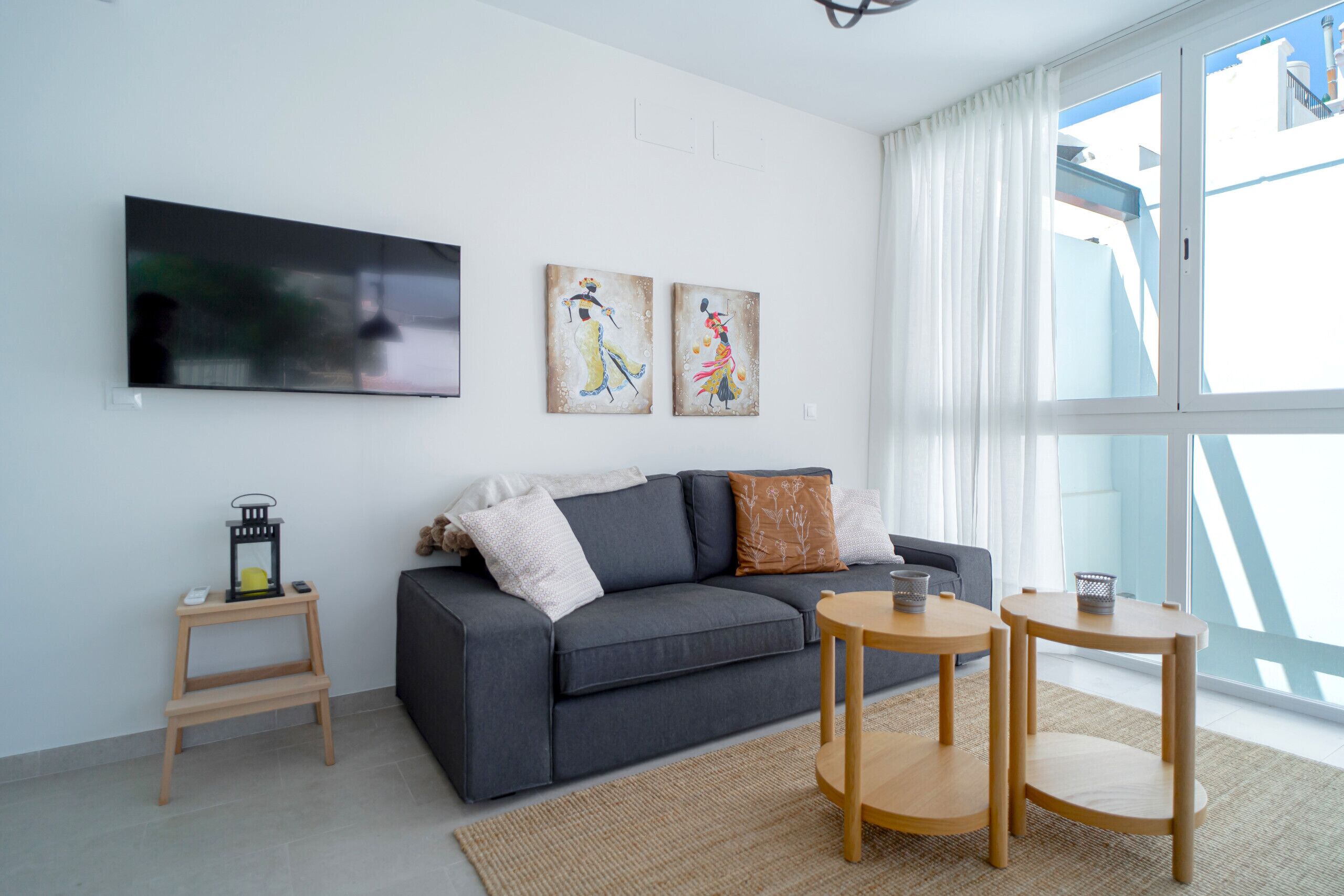 Un salon moderne avec un canapé gris, deux tables d’appoint en bois, un téléviseur à écran plat et deux œuvres d’art accrochées au mur.