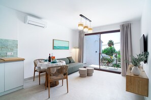 Ein modernes Wohnzimmer mit einer Couch, einem Esstisch und Stühlen. Ein Fernseher ist an der Wand montiert, ein Fenster mit Vorhängen und ein Balkon mit Gartenmöbeln vorhanden.