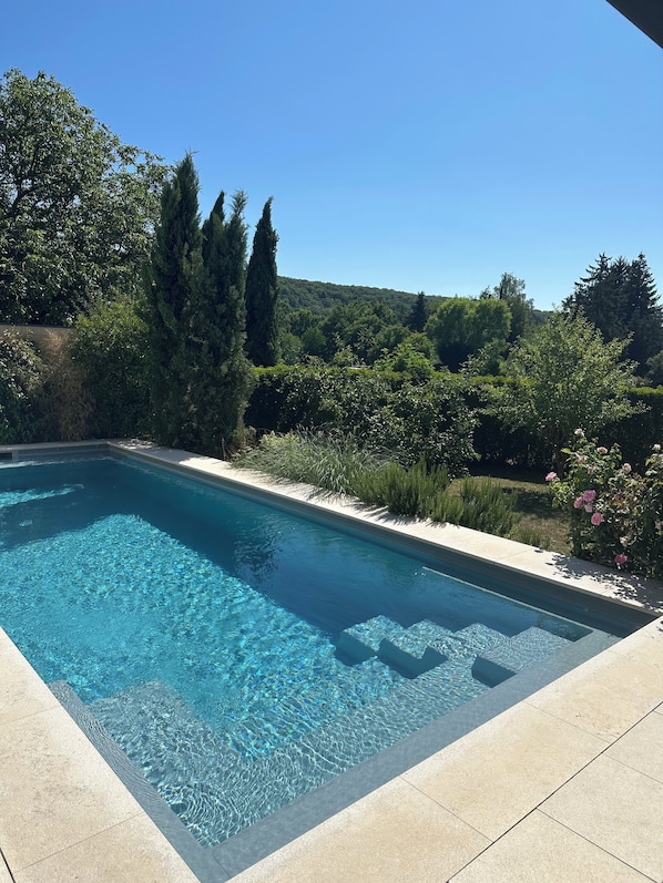 A rectangular swimming pool with clear blue water, surrounded by a tiled deck and lush greenery.