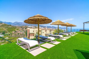 A row of sun loungers with thatched umbrellas on a grassy lawn, overlooking a coastal town and mountains. - Bayview Hills Suite by Casasol (Torrox)