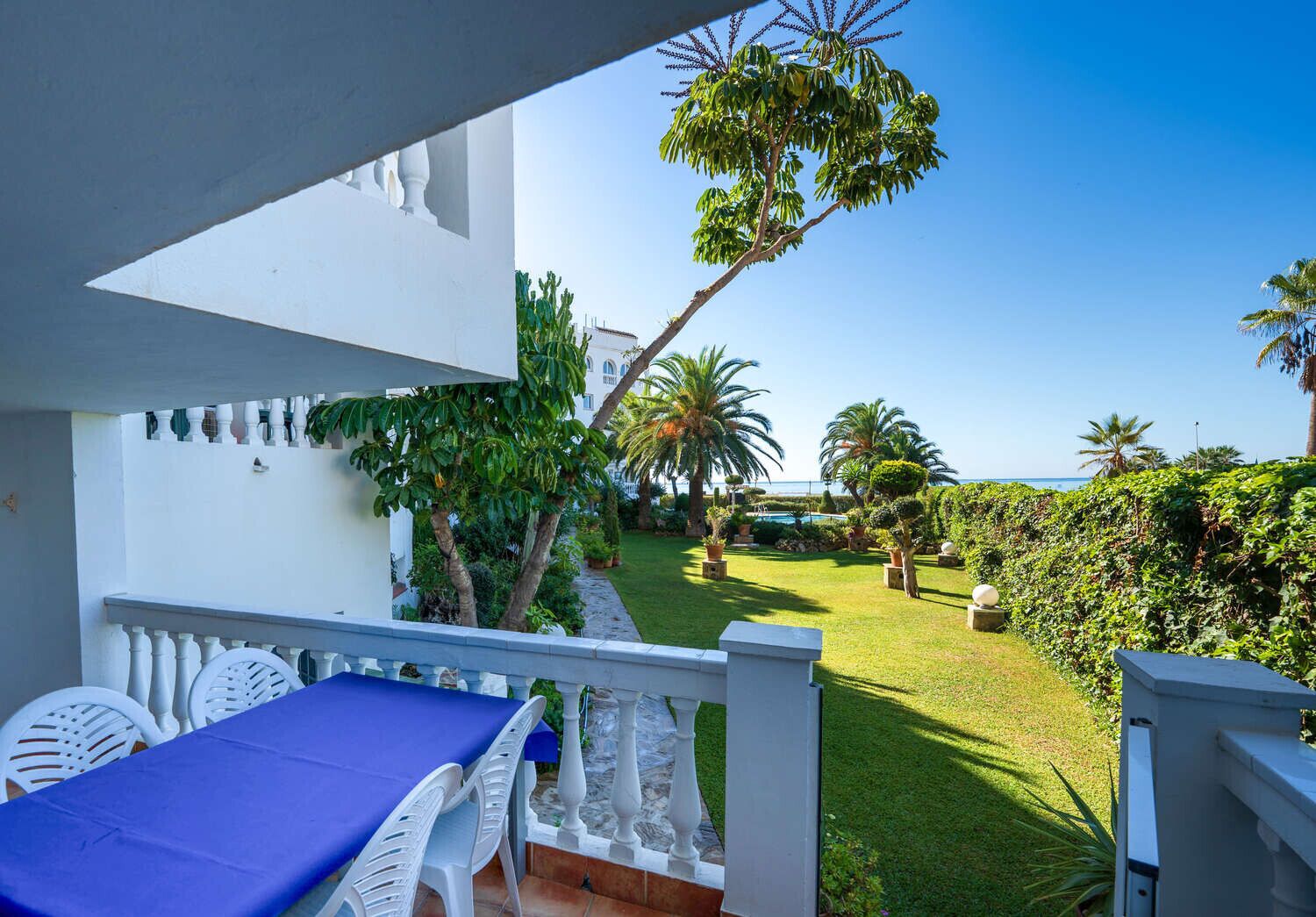 A balcony with a table and chairs, overlooking a well-maintained garden with various plants and a view of the sea.