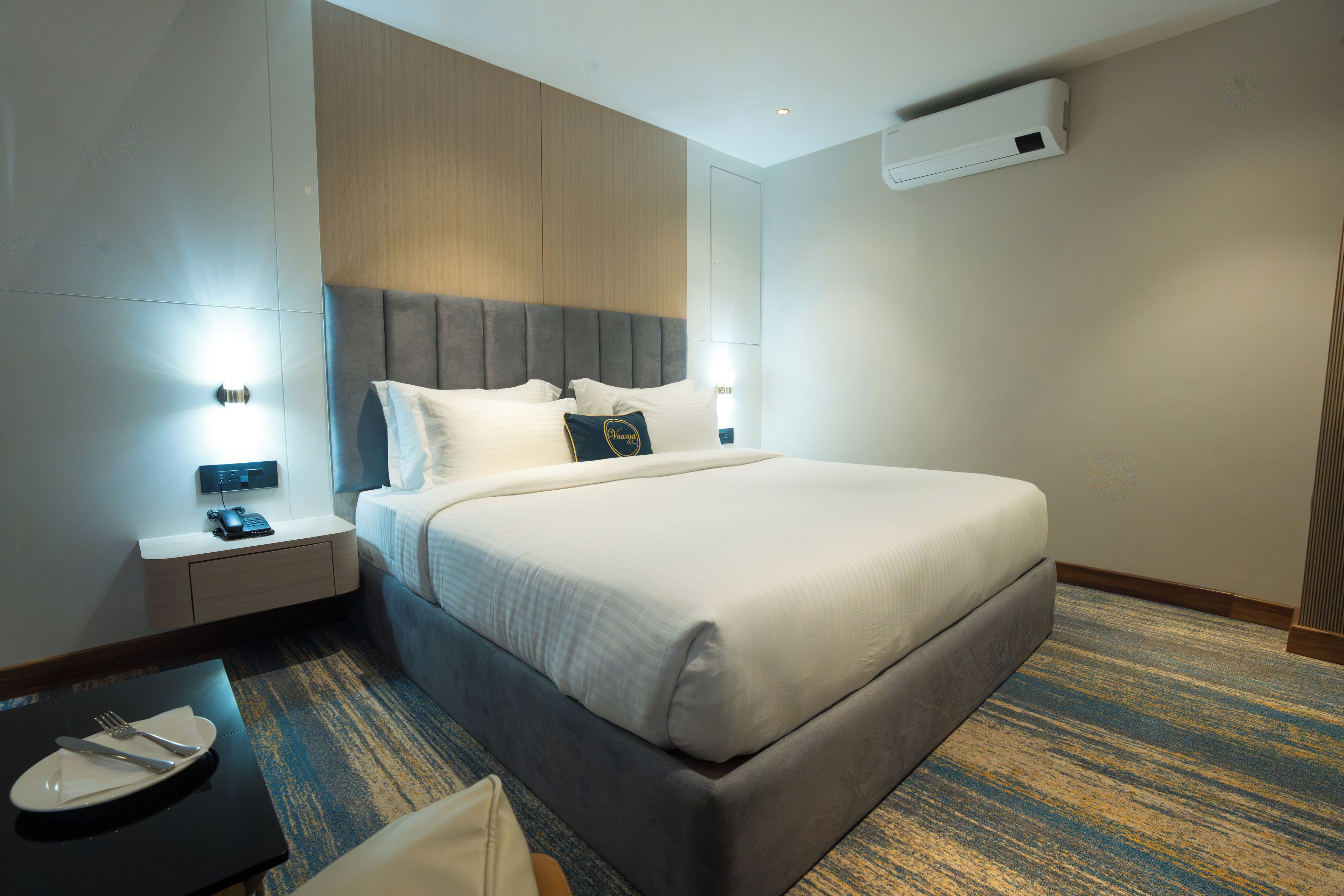Una habitación de hotel con una cama grande, mesitas de noche, un escritorio con teléfono y un equipo de aire acondicionado en la pared.