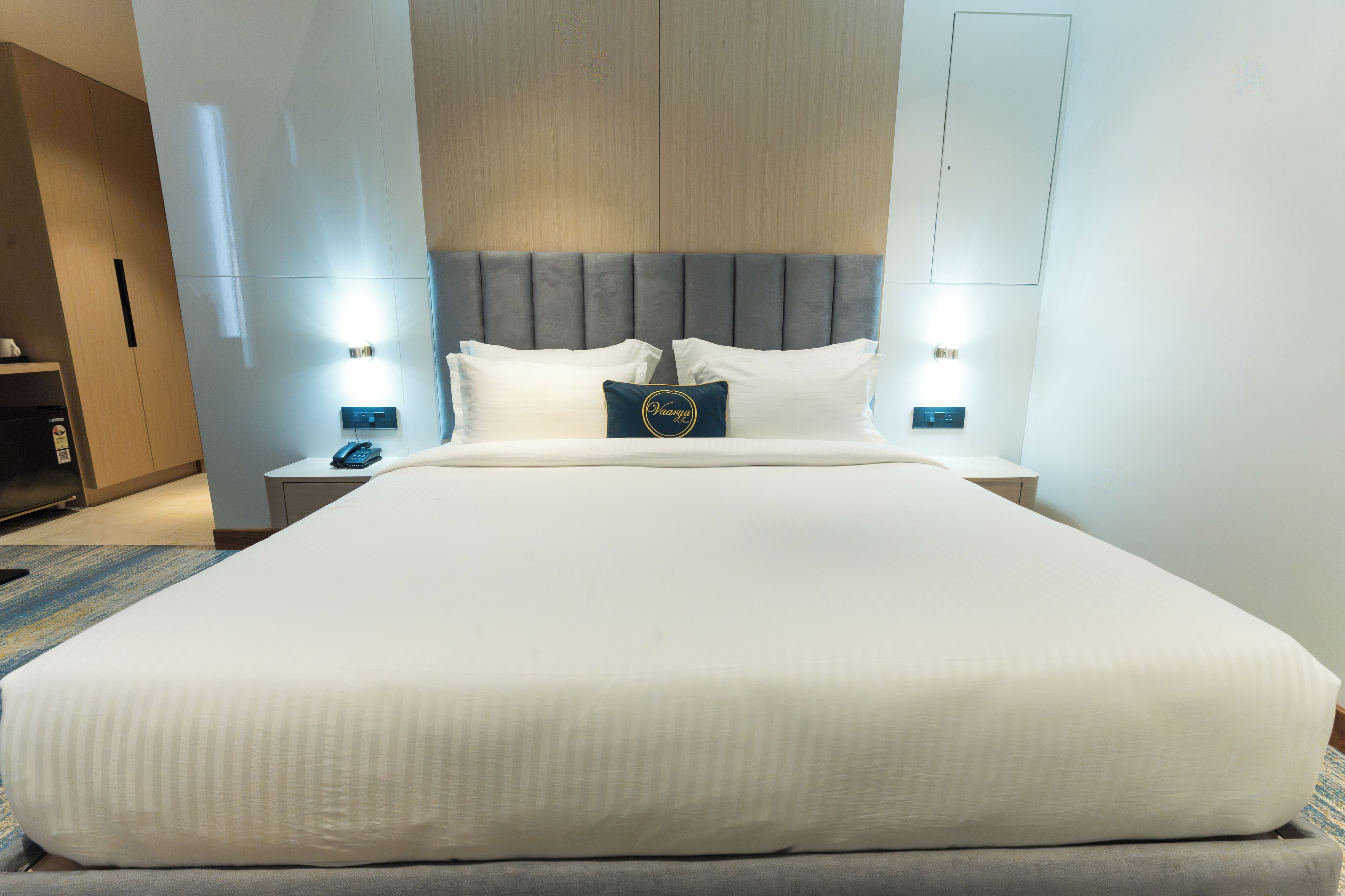 Uma cama grande com cabeceira estofada cinza, duas mesas de cabeceira com luminárias e um telefone pequeno na mesinha de cabeceira.