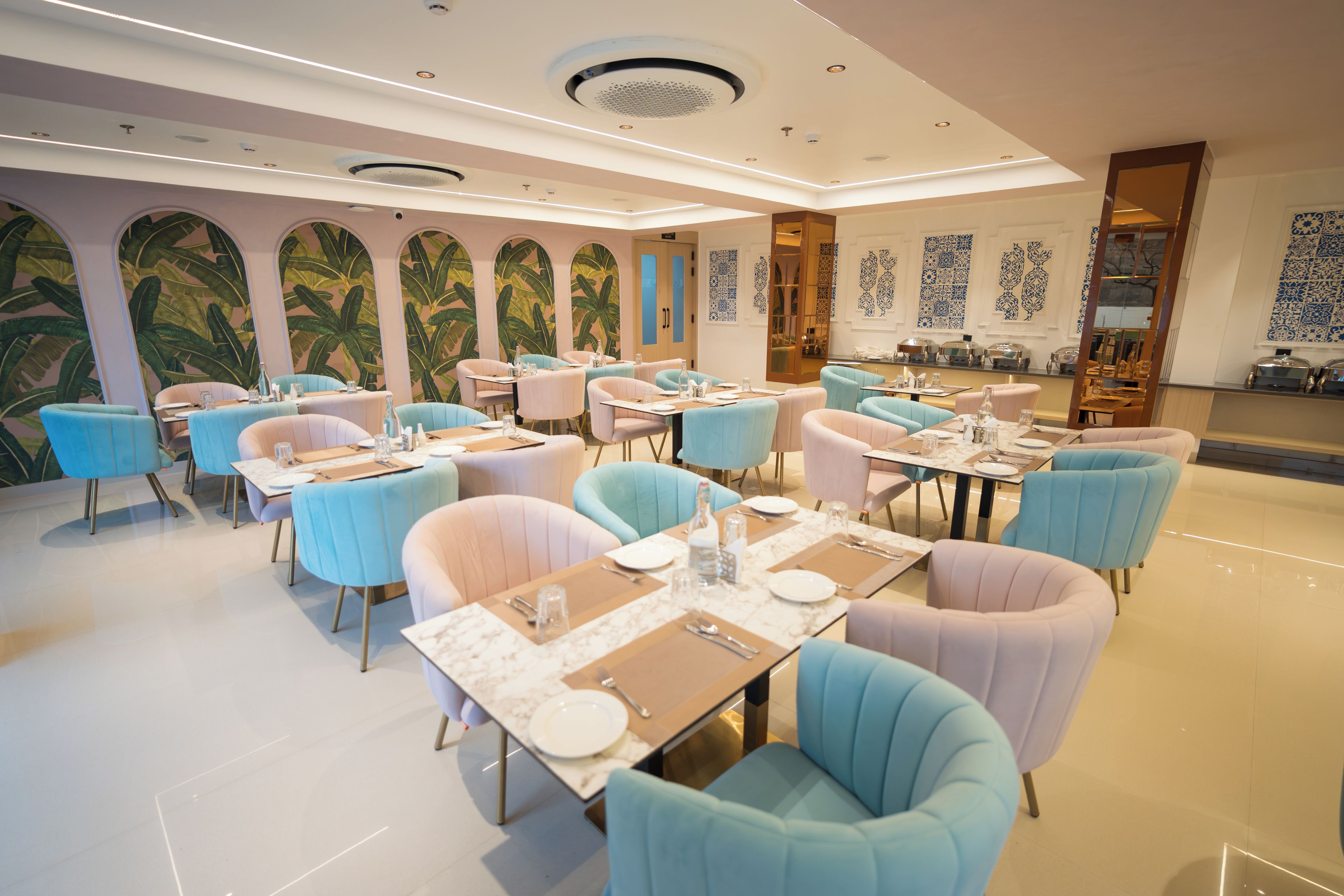 Un restaurante con varias mesas y sillas, un techo con iluminación empotrada y una pared con estampado de hojas tropicales.