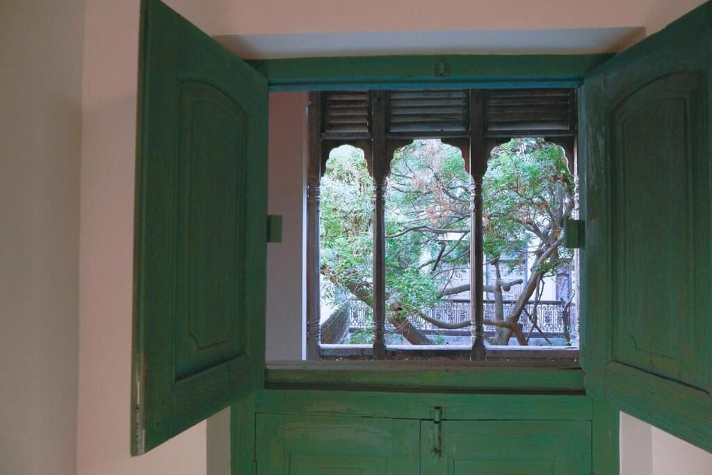 Una habitació amb una finestra i persianes verdes, vista a arbres i una paret blanca.