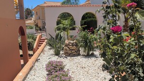 A courtyard with a white gravel ground, a stone wall, and various plants including a pink flowering bush. - Bright apartment with a spacious terrace near the ocean. (Mazarrón)