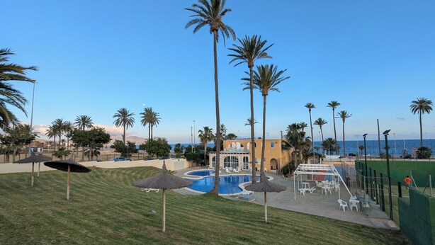 A pool area with palm trees, a building, and a view of the sea. - Bright apartment with a spacious terrace near the ocean. (Mazarrón)