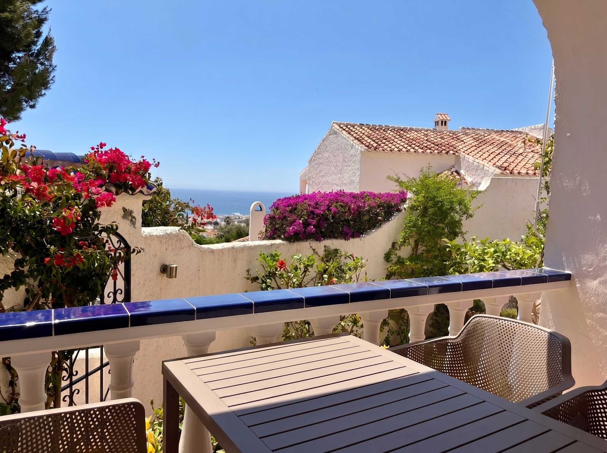 Een balkon met een tafel en stoelen, met uitzicht op de kust, gebouwen en bougainvillea.