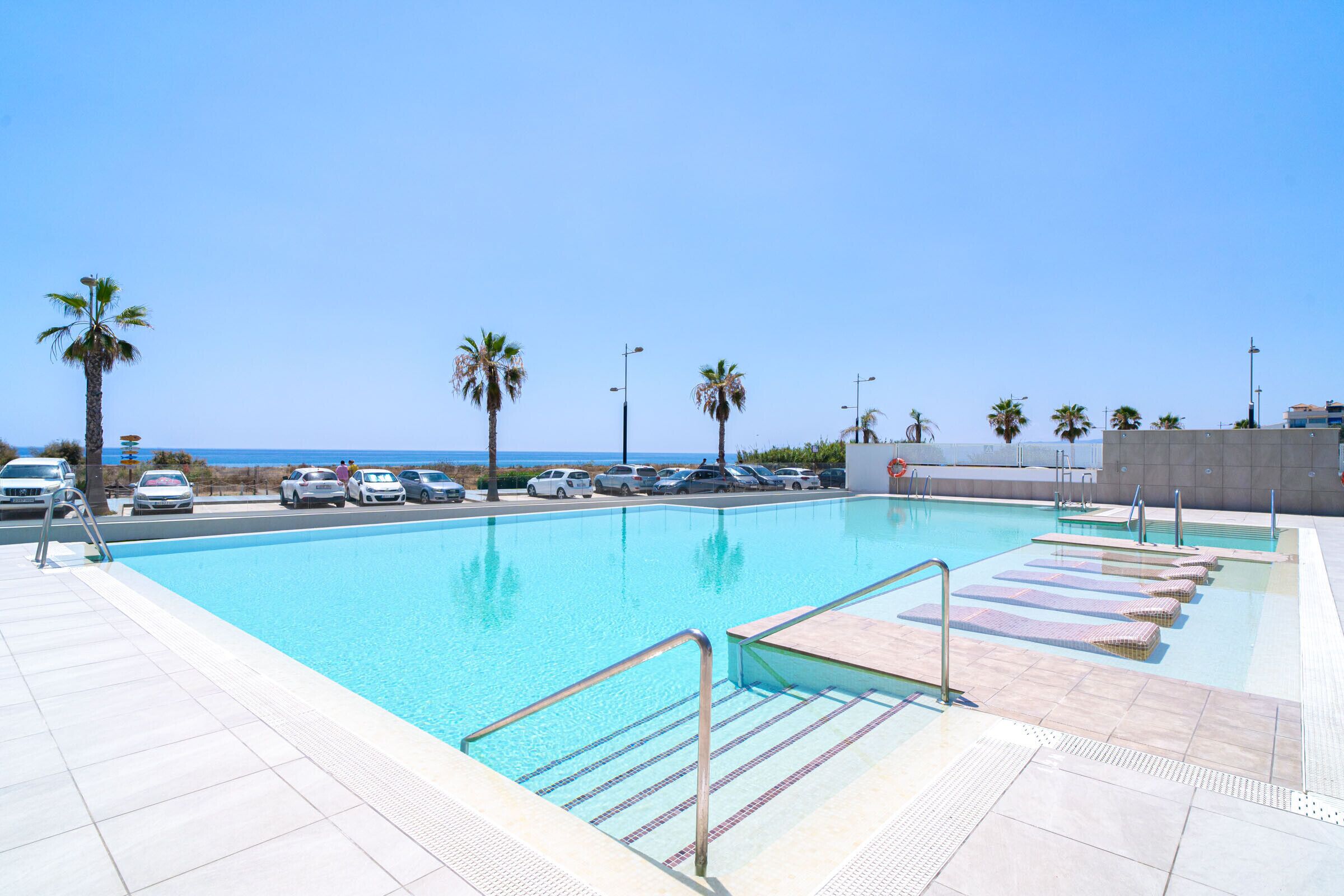 A swimming pool with a view of the sea, a parking area, and palm trees.