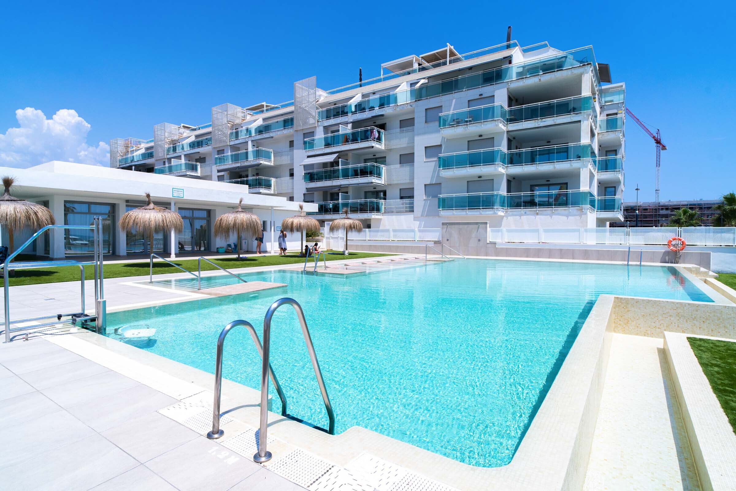 A modern hotel with a swimming pool, multiple balconies, and a clear blue sky.