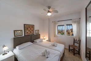 Ein Hotelzimmer mit zwei Betten, einem Deckenventilator, einem Fenster mit Vorhängen und gerahmten Bildern an der Wand.