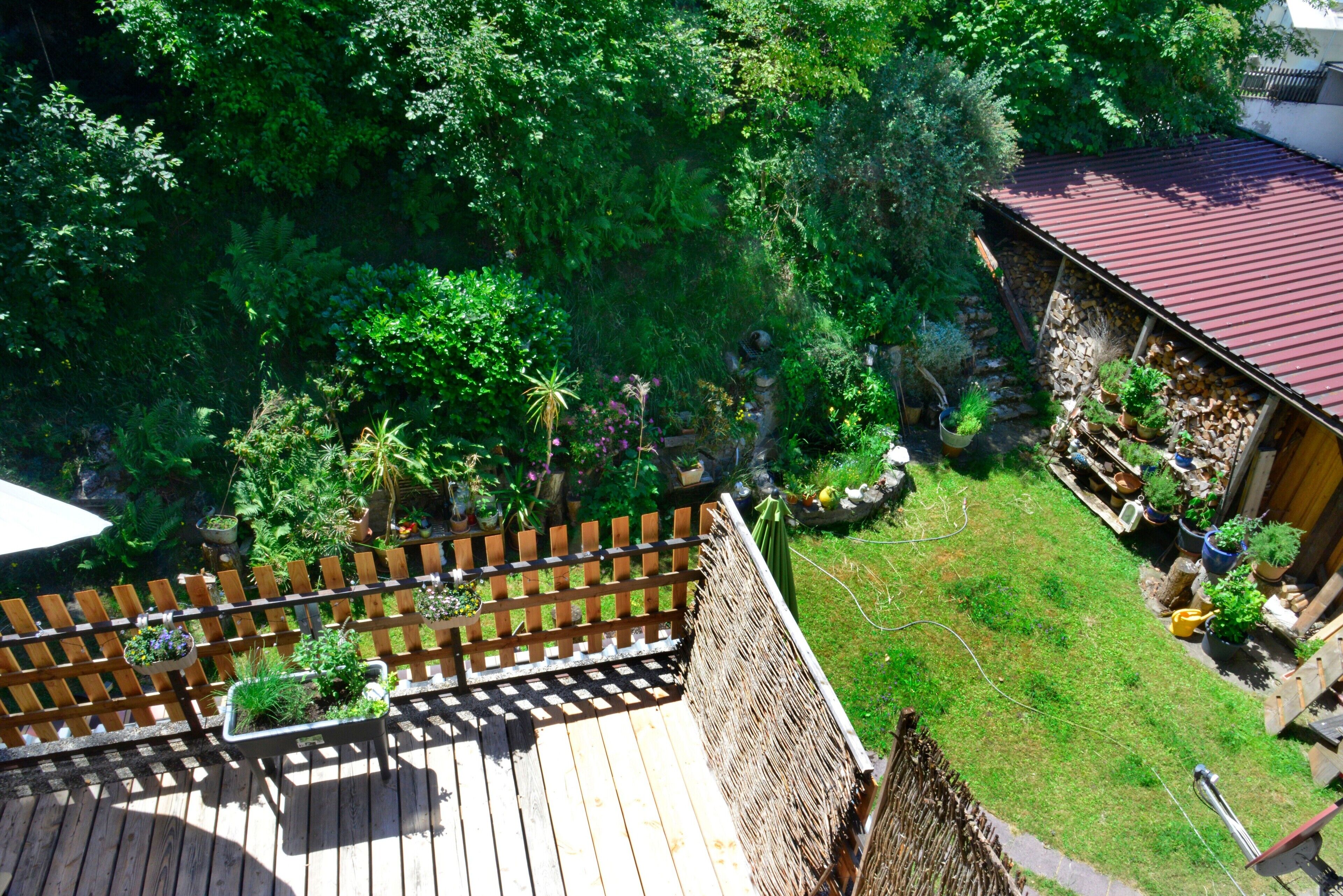 A wooden deck with a wooden fence, a garden with various plants, and a building with a red roof.