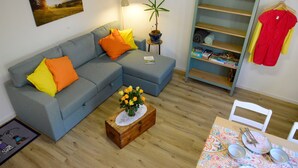 Un salon avec un canapé gris, des coussins orange, une petite table sur laquelle est posé un vase de fleurs, une plante en pot et un coin repas avec une table dressée pour deux personnes.