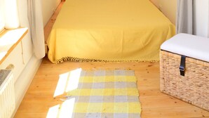 Une chambre à coucher avec un lit, des rideaux, une table de chevet, une lampe, un panier et un tapis à carreaux.