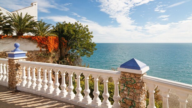 Un balcón costero con barandilla blanca, pared de piedra y vista al mar.