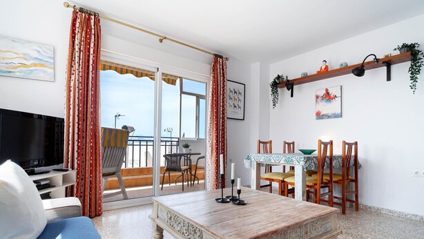 Ein Wohnzimmer mit Fernseher, einer Couch, einem Couchtisch, einem Essbereich mit Stühlen und einem Balkon mit Tisch. Zusätzlich befindet sich ein Fernseher auf dem Balkon.
