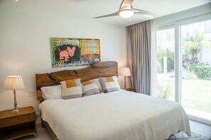 A hotel room with a bed, a ceiling fan, a nightstand, a lamp, a window with curtains, and a colorful artwork on the wall. - Beautiful Ground Floor Condo with Spectacular Ocean Views and Resort Style Amenities (Playa Tamarindo, Guanacaste Province)