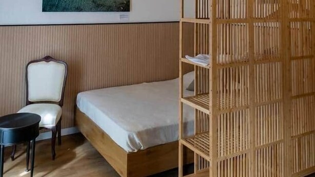 Een kamer met een bed, een stoel, een houten plankunit en een schilderij aan de muur.