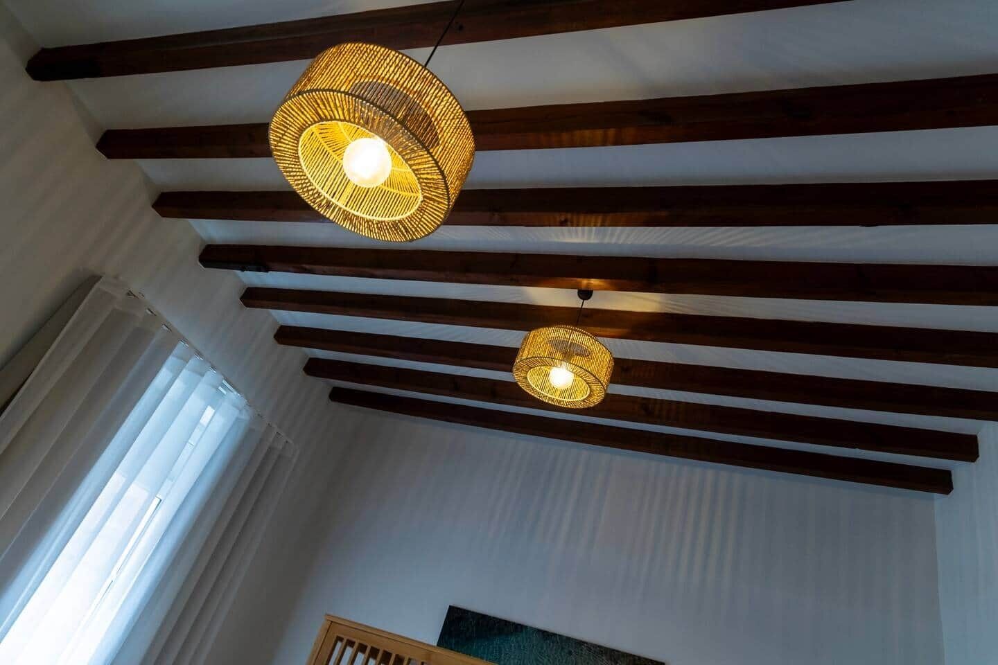 Quarto com vigas de madeira aparentes, luminária de fibra natural e janela com persianas.