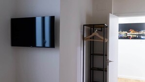 Una stanza con una TV a schermo piatto fissata al muro, una gruccia di legno su un porta appendiabiti in metallo nero, e una porta che dà su uno spazio esterno con vista sulla città.