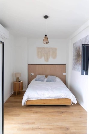 A bedroom with a wooden bed, a hanging light fixture, a small bedside table, and a wall-mounted artwork. - Suite 5 Apartment, 24 Teatro (Alicante)
