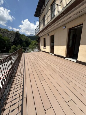 A wooden deck with a metal railing, a building with a balcony, and a mountainous backdrop.