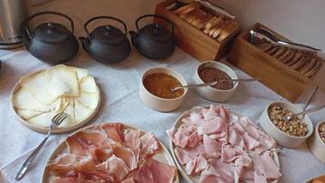 Une table sur laquelle sont disposés divers aliments et ustensiles, notamment des assiettes de jambon et de fromage en tranches, des bols de condiments et une boîte à pain en bois.