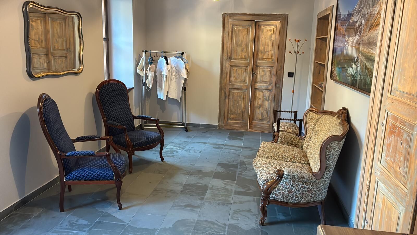 Una habitación con zona de estar que incluye dos sillones y un sofá, un perchero con prendas de vestir, una puerta de madera y un cuadro en la pared.
