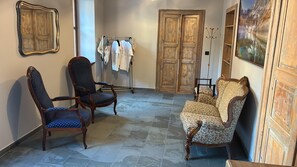 A room with a seating area featuring two armchairs and a sofa, a clothing rack with garments, a wooden door, and a painting on the wall.
