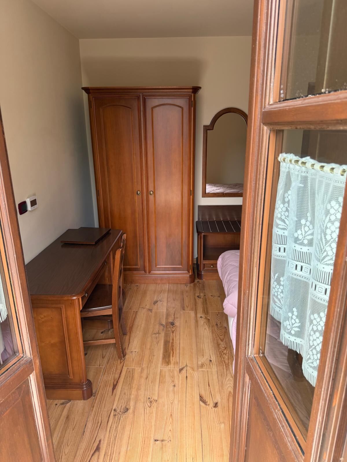 A bedroom with a wooden wardrobe, a bedside table, a mirror, and a window with lace curtains.